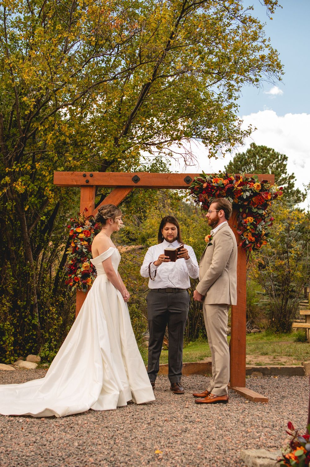 Couple exchanges vows under rustic wooden arch adorned with fall florals during outdoor autumn wedding ceremony.