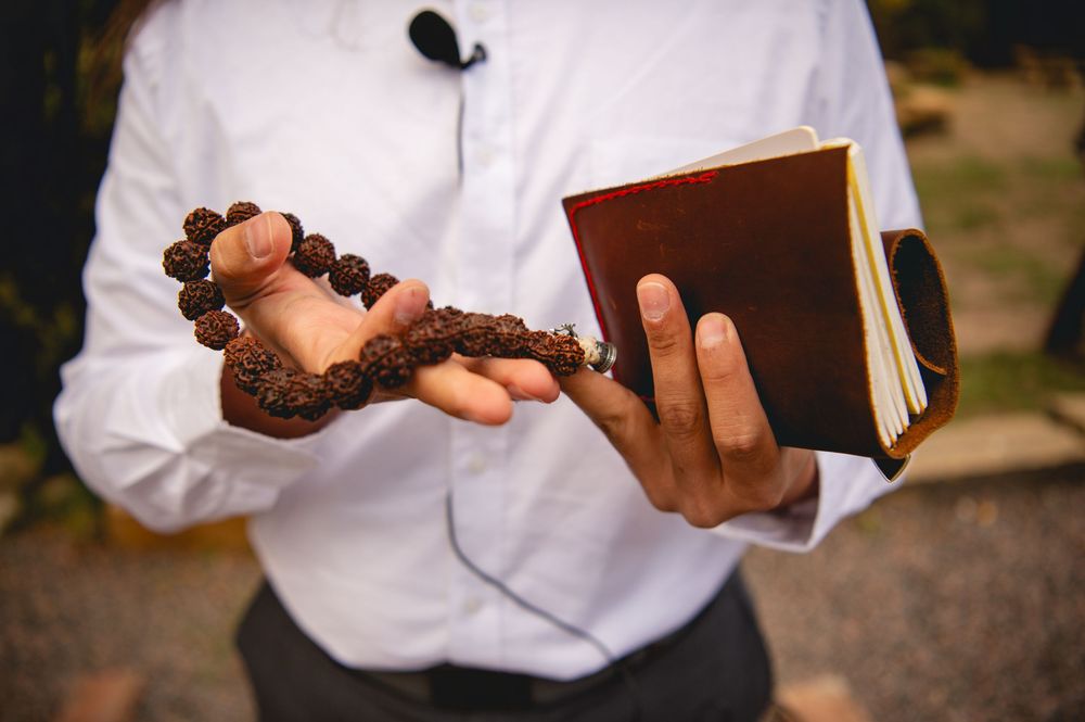Person holding wooden prayer beads and a brown leather book outdoors, wearing a white shirt.