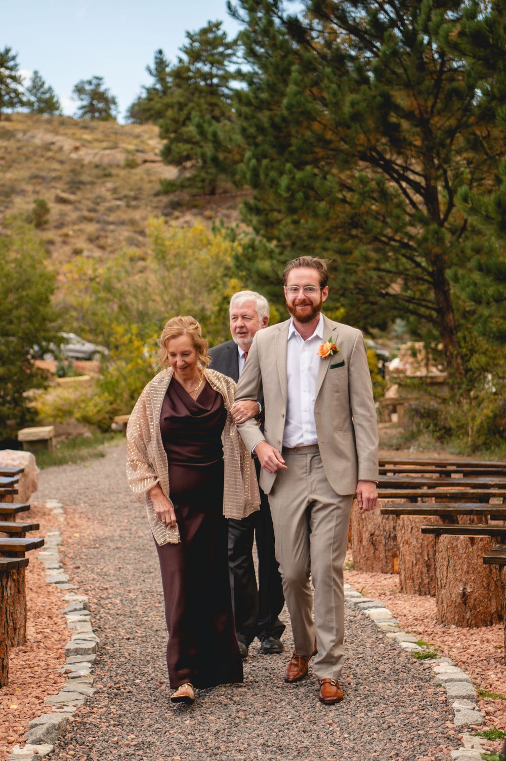 Groom in tan suit walks with parents down outdoor wedding aisle surrounded by pine trees and rustic wooden benches.
