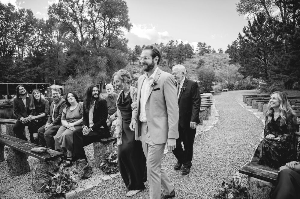 Groom in tan suit walks down outdoor wedding aisle as guests watch from rustic wooden benches.