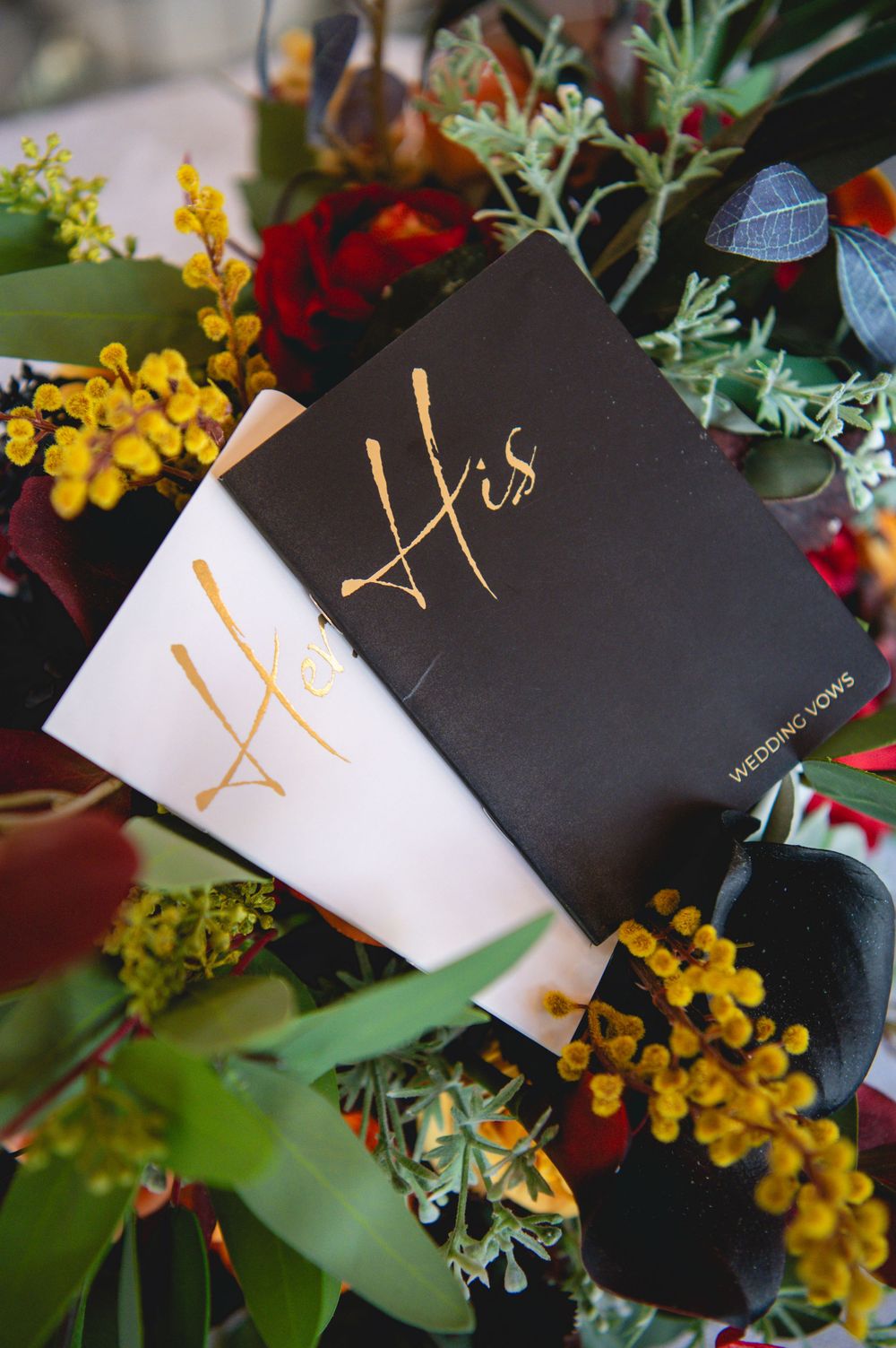His and Hers gold calligraphy wedding vow books in black and white nestled among red roses and yellow wildflowers.