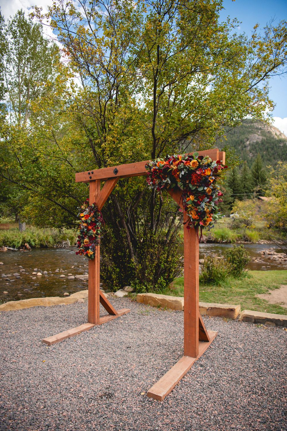 Rustic wooden wedding arch adorned with colorful fall flowers, set on gravel near a mountain stream with autumn trees.