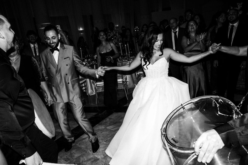 Black and white photo of bride and groom dancing joyfully at wedding reception surrounded by guests.