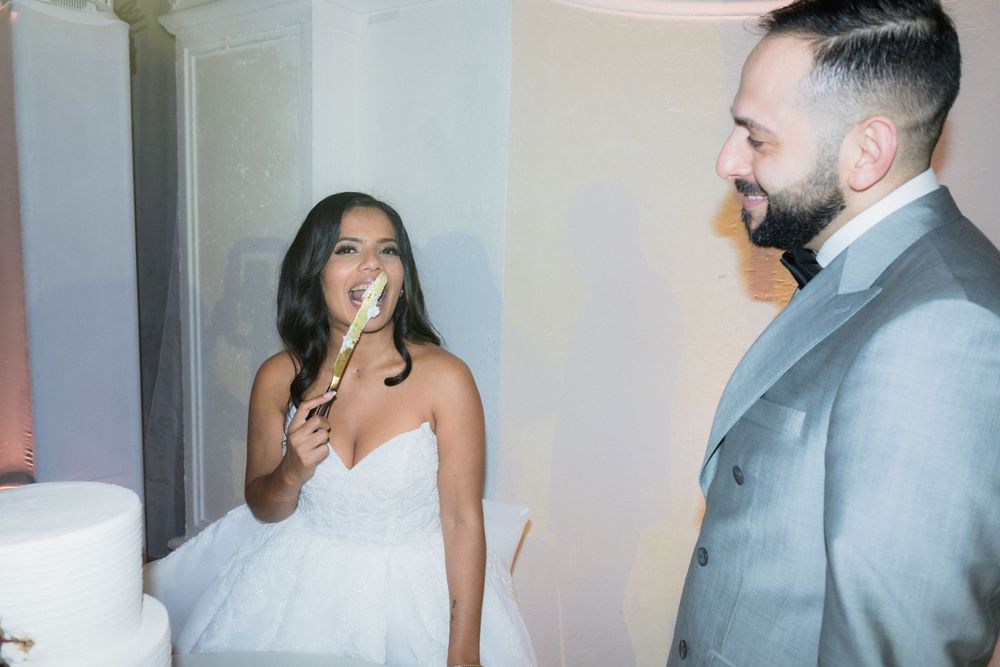 Bride in white strapless gown licking cake knife beside groom in gray suit at wedding reception.