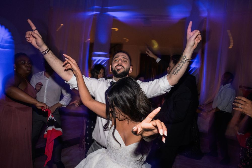 Bride and groom dancing joyfully with guests at wedding reception under blue lights.
