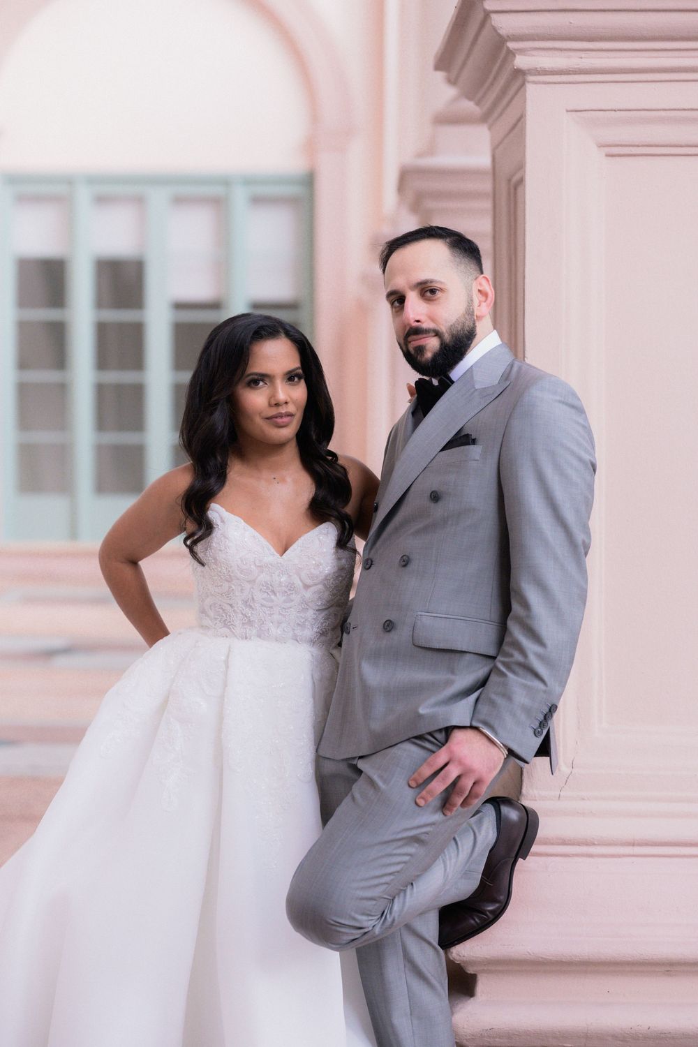 Bride in white gown and groom in gray suit pose elegantly against a pink architectural backdrop.