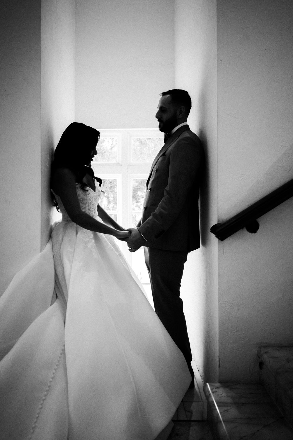 Black and white photo of bride and groom holding hands on staircase, sharing a romantic moment in wedding attire.