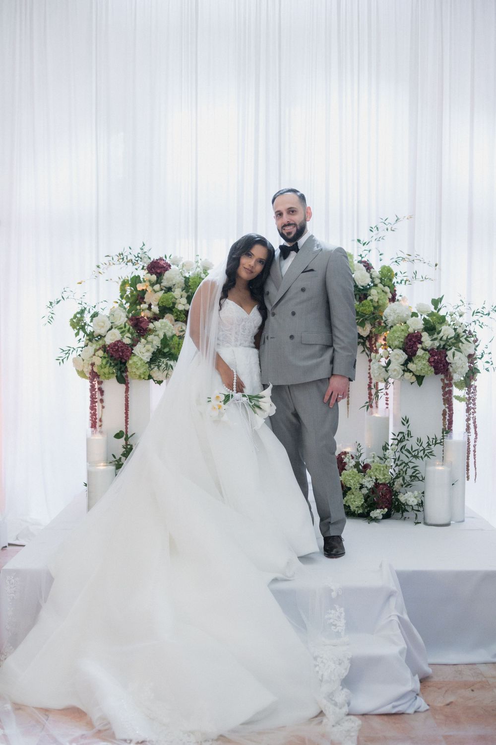 Bride in white ball gown and groom in gray suit pose together at elegant floral wedding altar with white draping.