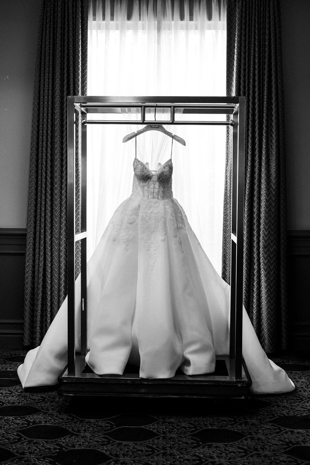 Black and white photo of an elegant lace wedding gown displayed on a hanger inside a glass case by a window.