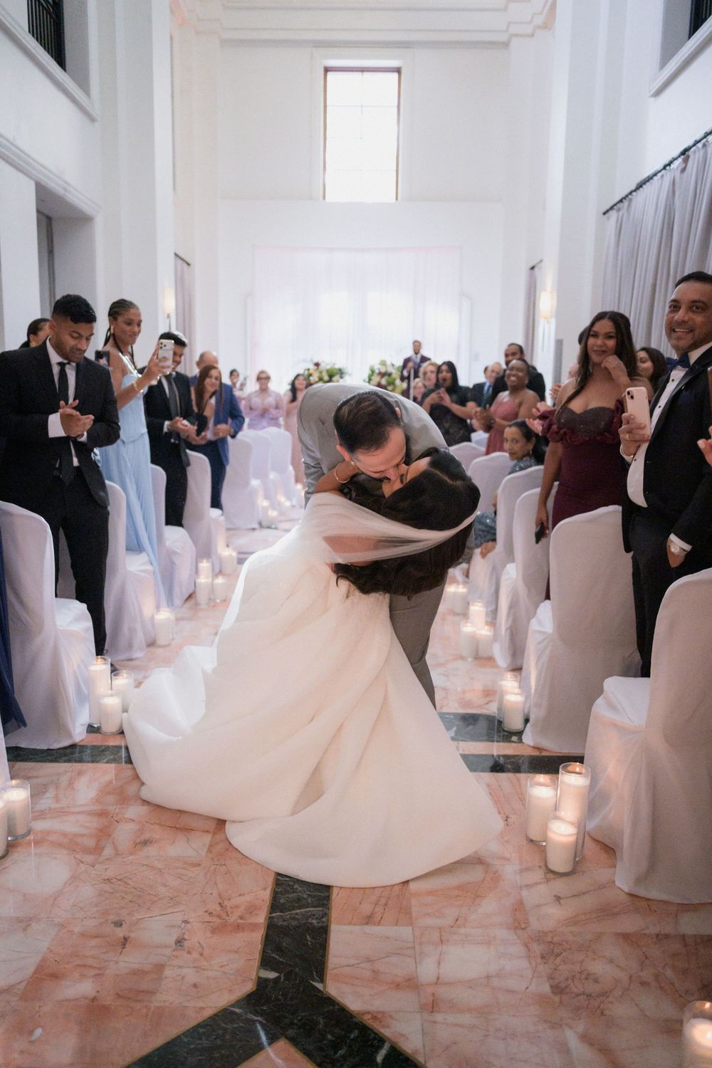 Groom dips bride for a romantic kiss at wedding ceremony aisle lined with candles and white chairs.