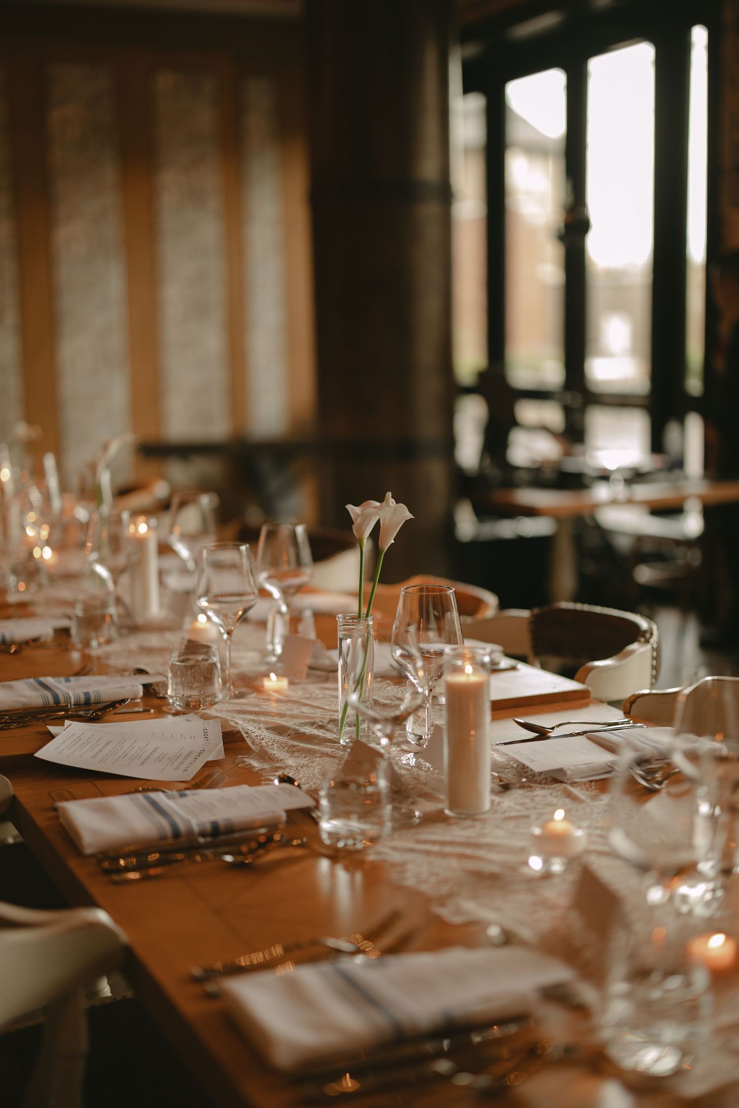 Long rustic wooden wedding reception table set with lace runners, white candles, calla lilies, and glassware.