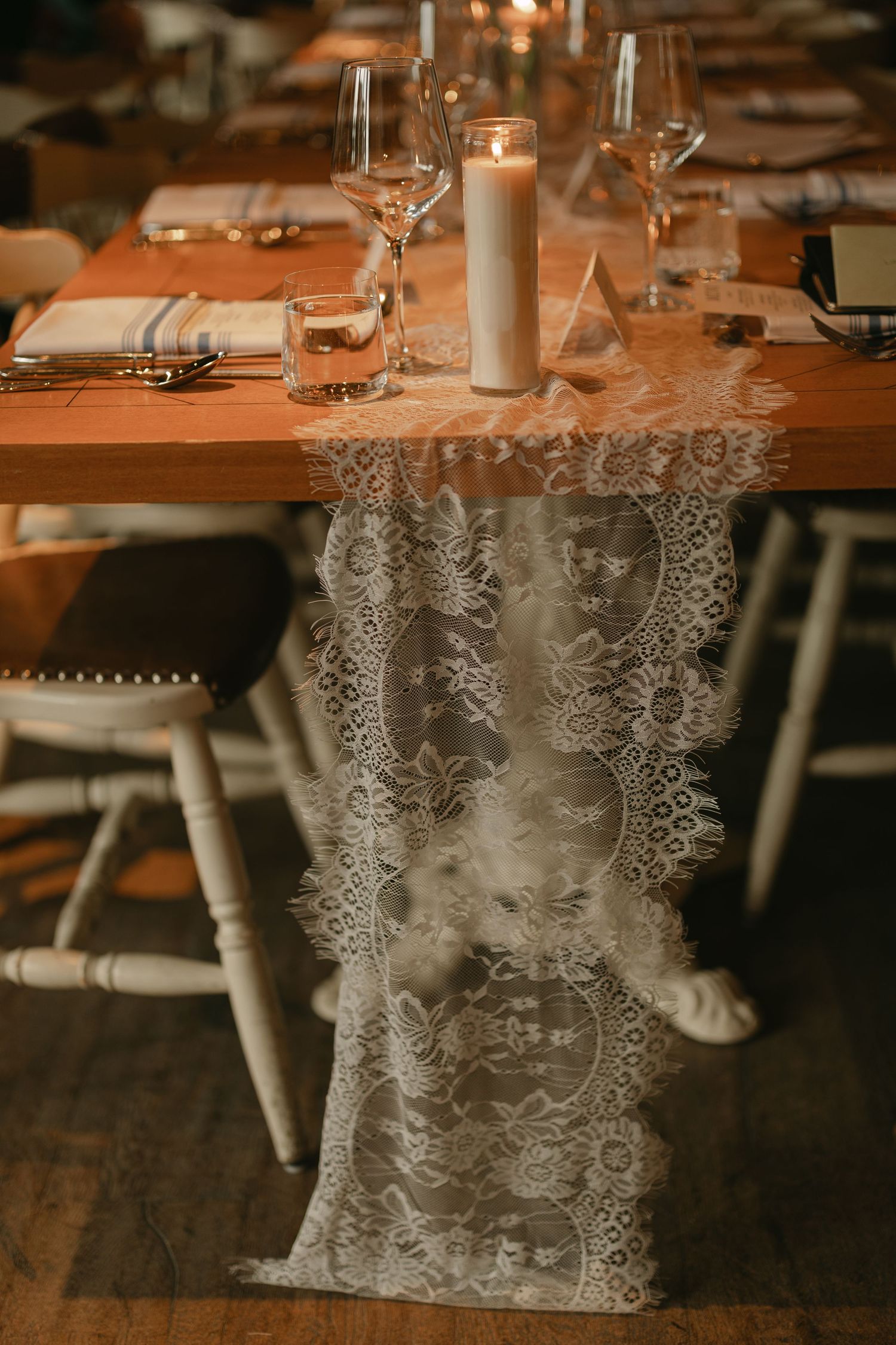 Rustic wooden reception table adorned with delicate lace runner, white pillar candle, and crystal glassware.