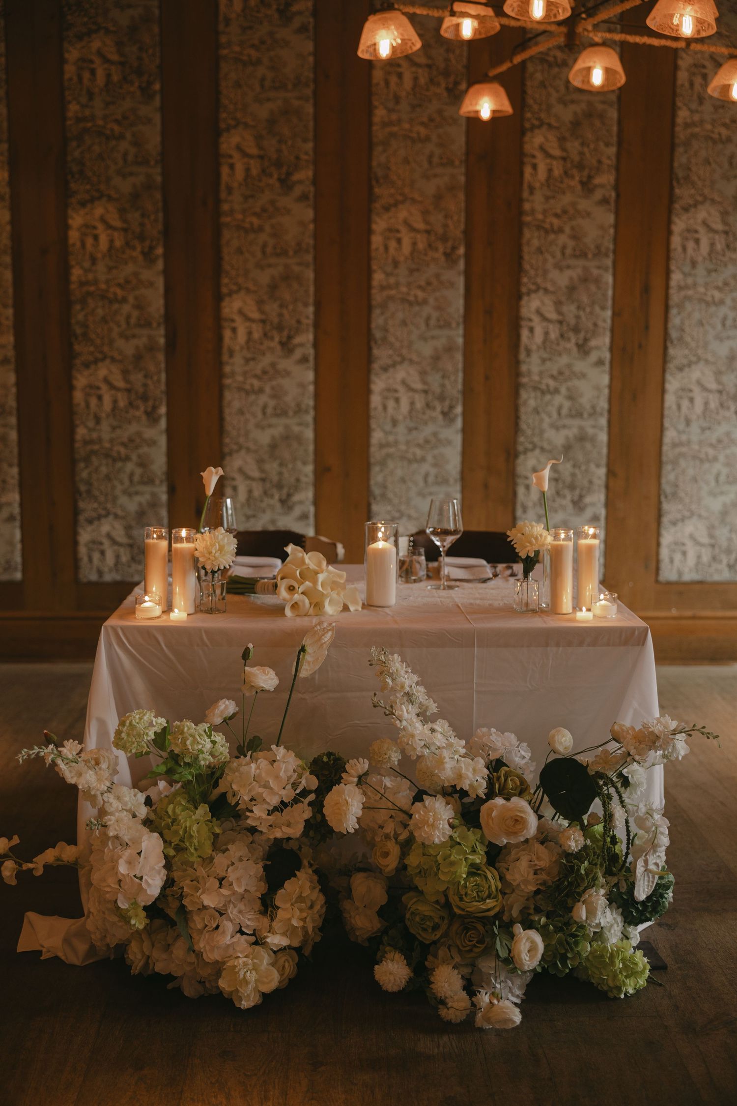 Elegant wedding sweetheart table with white floral arrangements, candles, and warm pendant lights in a decorated reception hall.