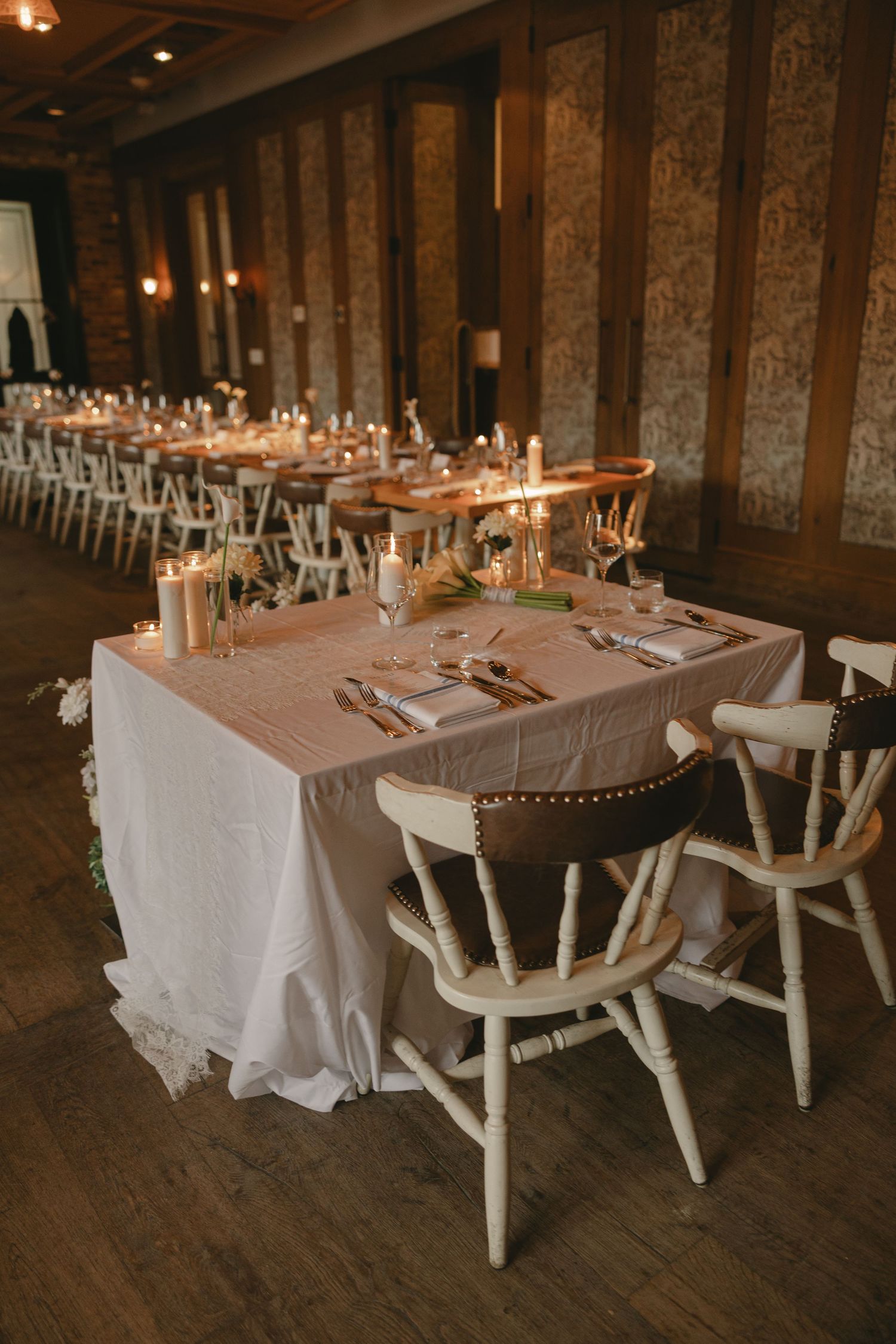Wedding reception tables with blush linen, candles, white crossback chairs, and warm candlelight in an elegant venue.