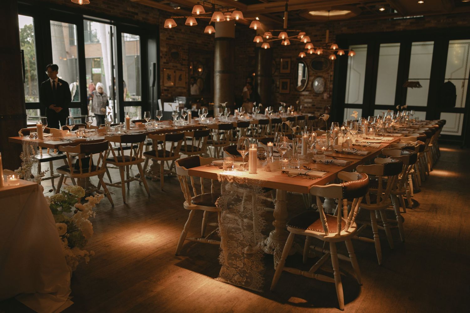 Rustic wedding reception venue with long wooden tables, white crossback chairs, and warm globe pendant lighting.