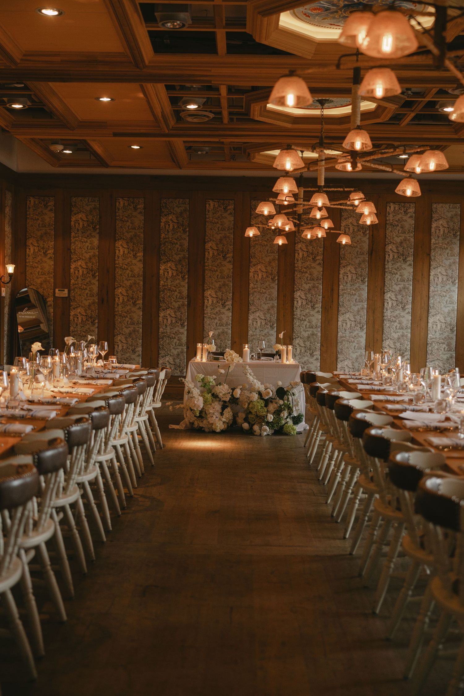 Grand wedding reception hall with long banquet tables, white chairs, floral centerpieces, and copper pendant chandeliers.