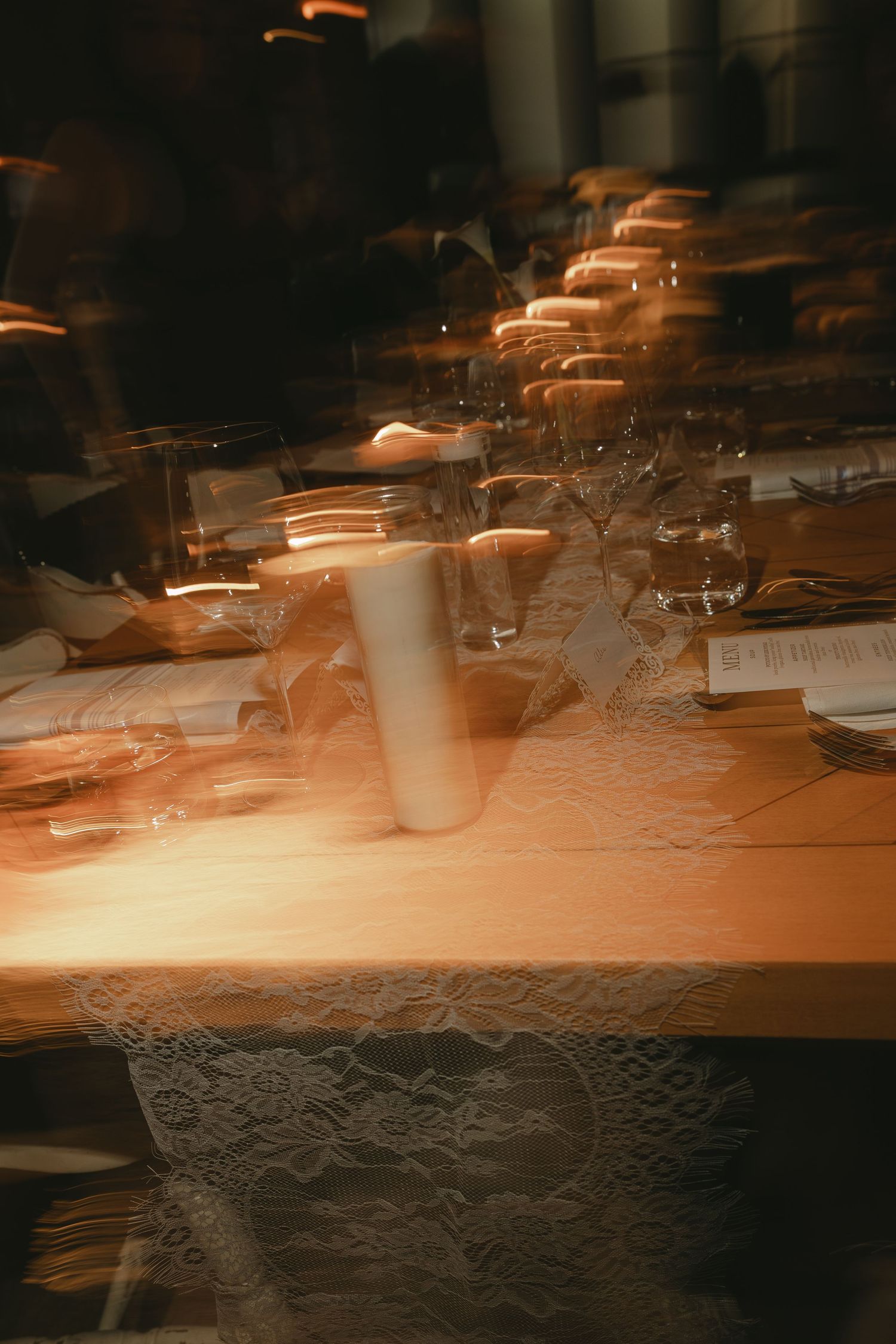 Blurred artistic shot of wedding reception table with lace runner, candles, and glassware in warm candlelight.