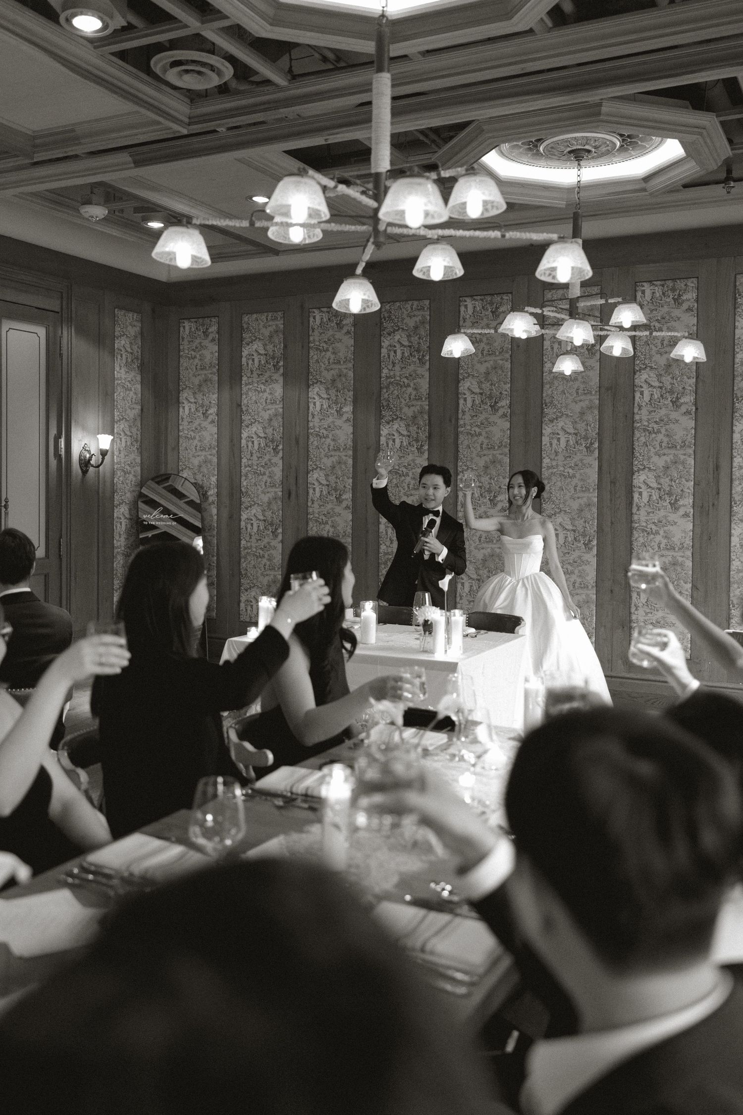 Black and white photo of wedding couple toasting with guests during reception in an elegant chandelier-lit ballroom.