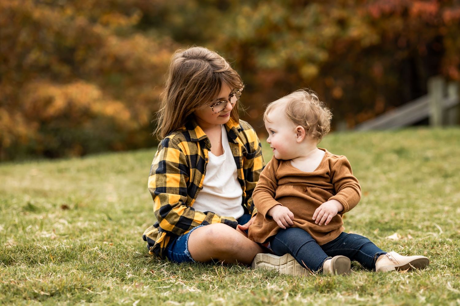 A young girl in a plaid shirt sits on grass with a toddler in autumn, sharing a sweet moment outdoors.