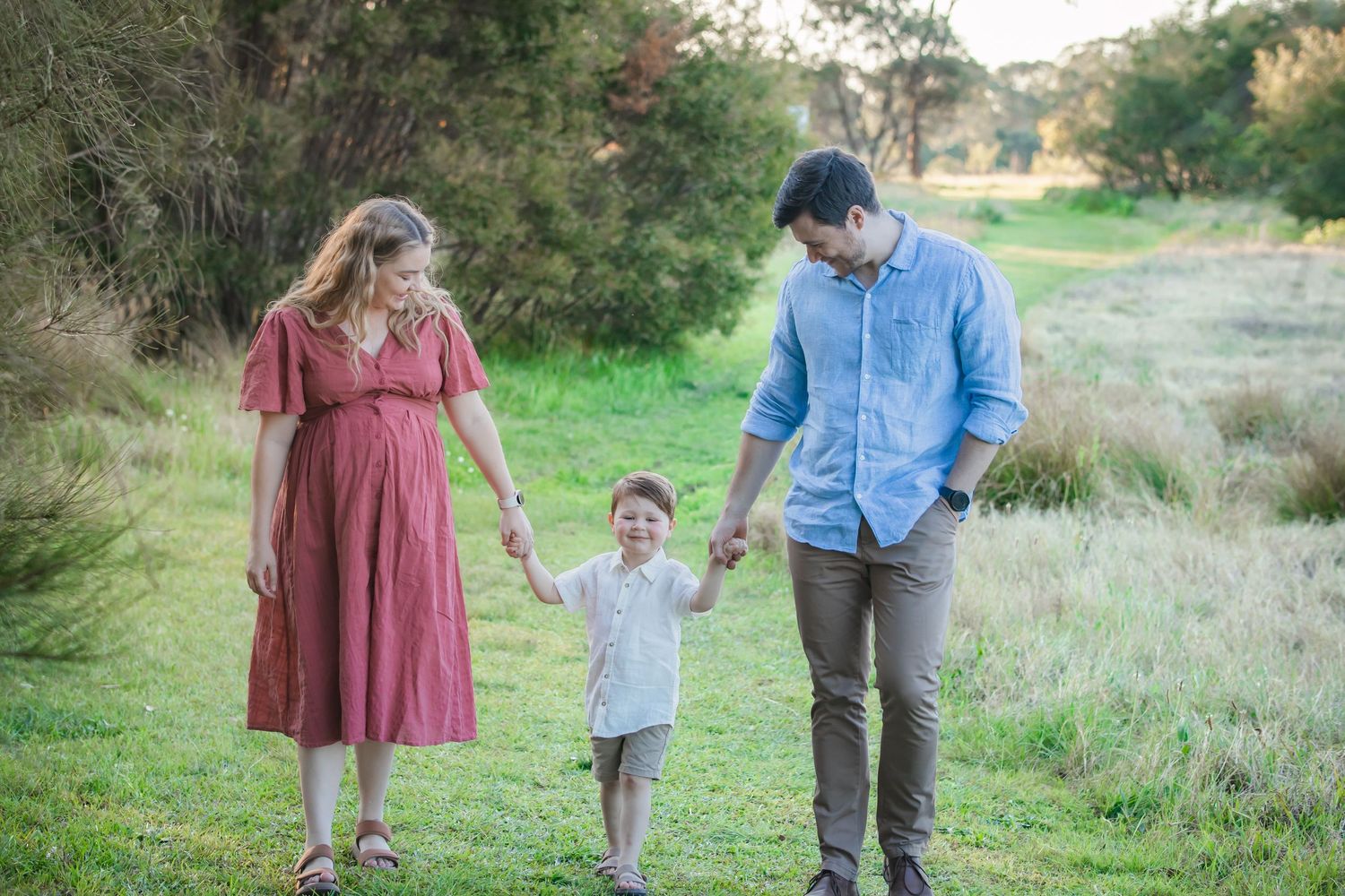 Pregnant woman in red dress and man in blue shirt hold hands with toddler son while walking in green field at Rouse Hill Regional Park.
