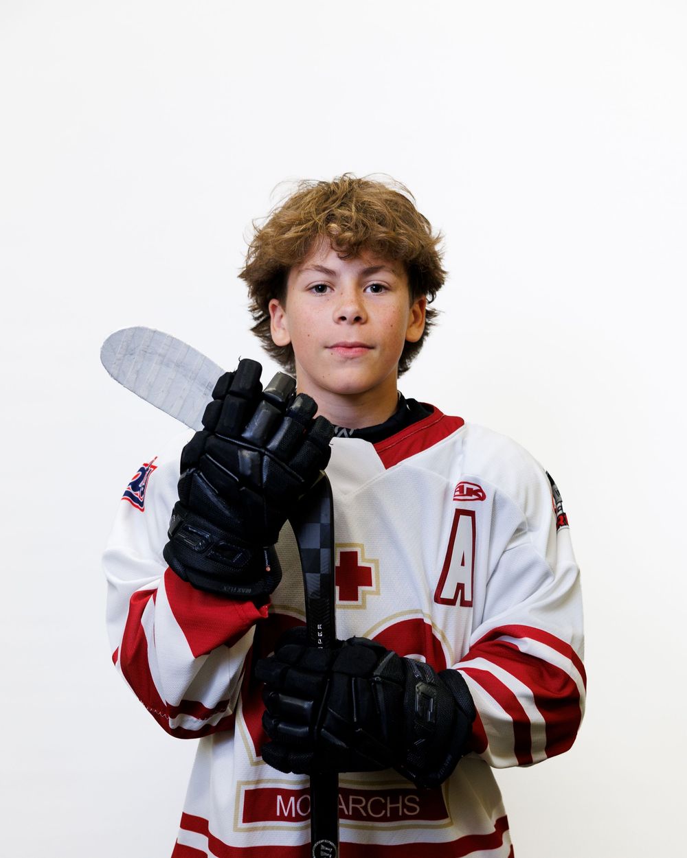 Young hockey player in white Monarchs jersey holding a stick, wearing black gloves with an A patch on uniform.