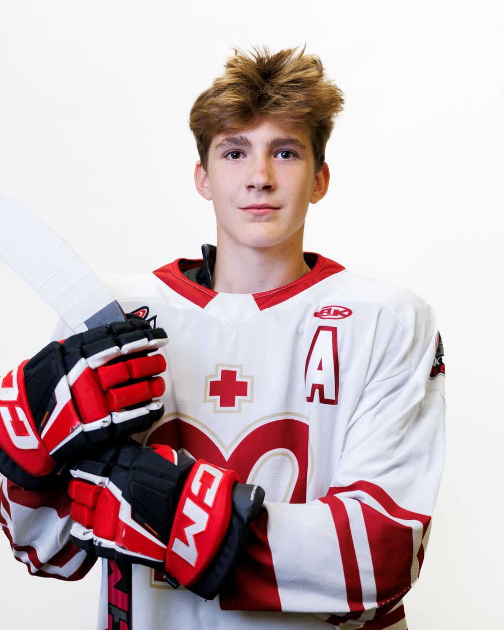 Young hockey player in white and red jersey with letter A patch, holding a stick, wearing black and red CCM gloves.