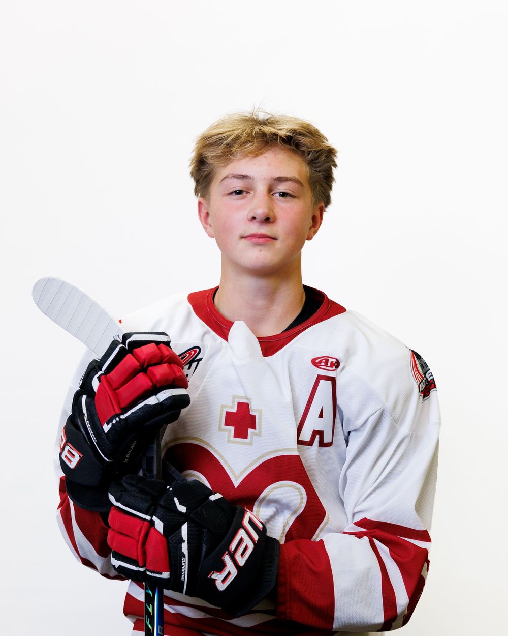 Young hockey player in white and red jersey holding a stick and Bauer gloves, posing for a team portrait photo.