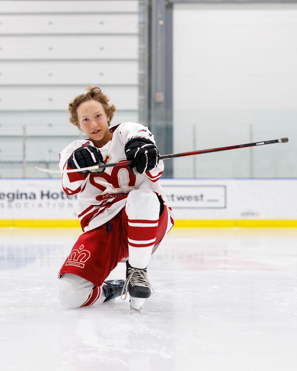 Young hockey player in red and white uniform kneeling on ice rink holding a stick during a game.