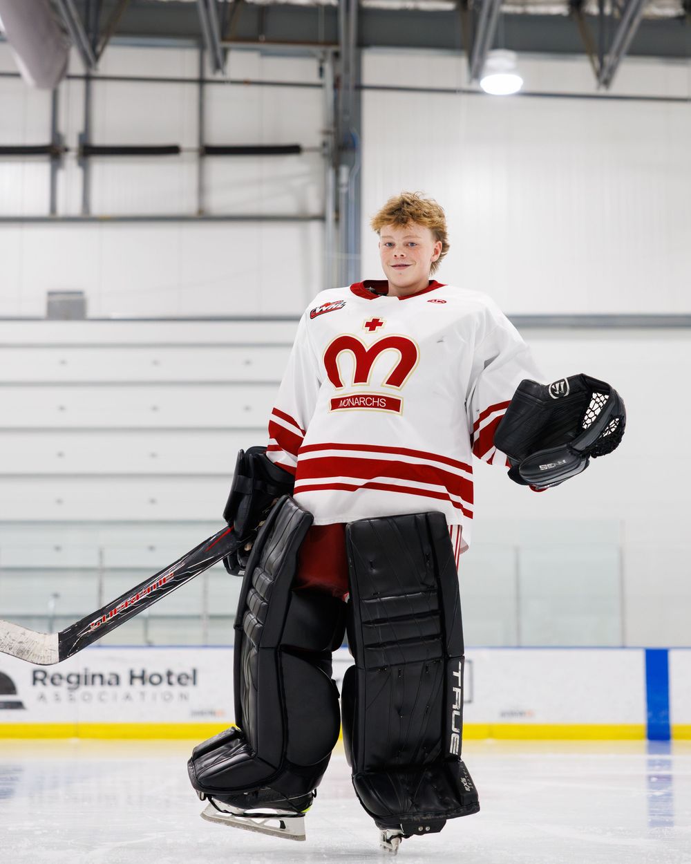 Young hockey goalie in white and red uniform poses on ice rink holding helmet at Regina Hotel Arena.