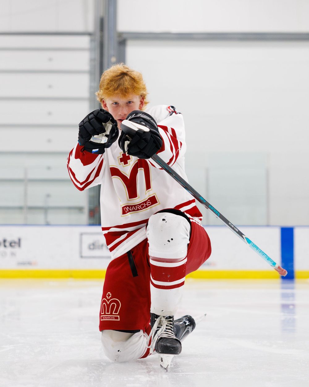 Young hockey player in white and red jersey kneeling on ice, holding a stick in a celebratory pose.