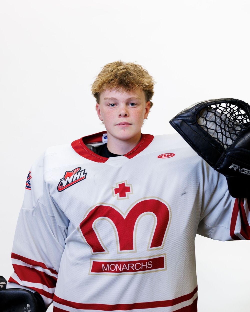 Young hockey player in white WHL Monarchs jersey holding goalie equipment during team photo session.
