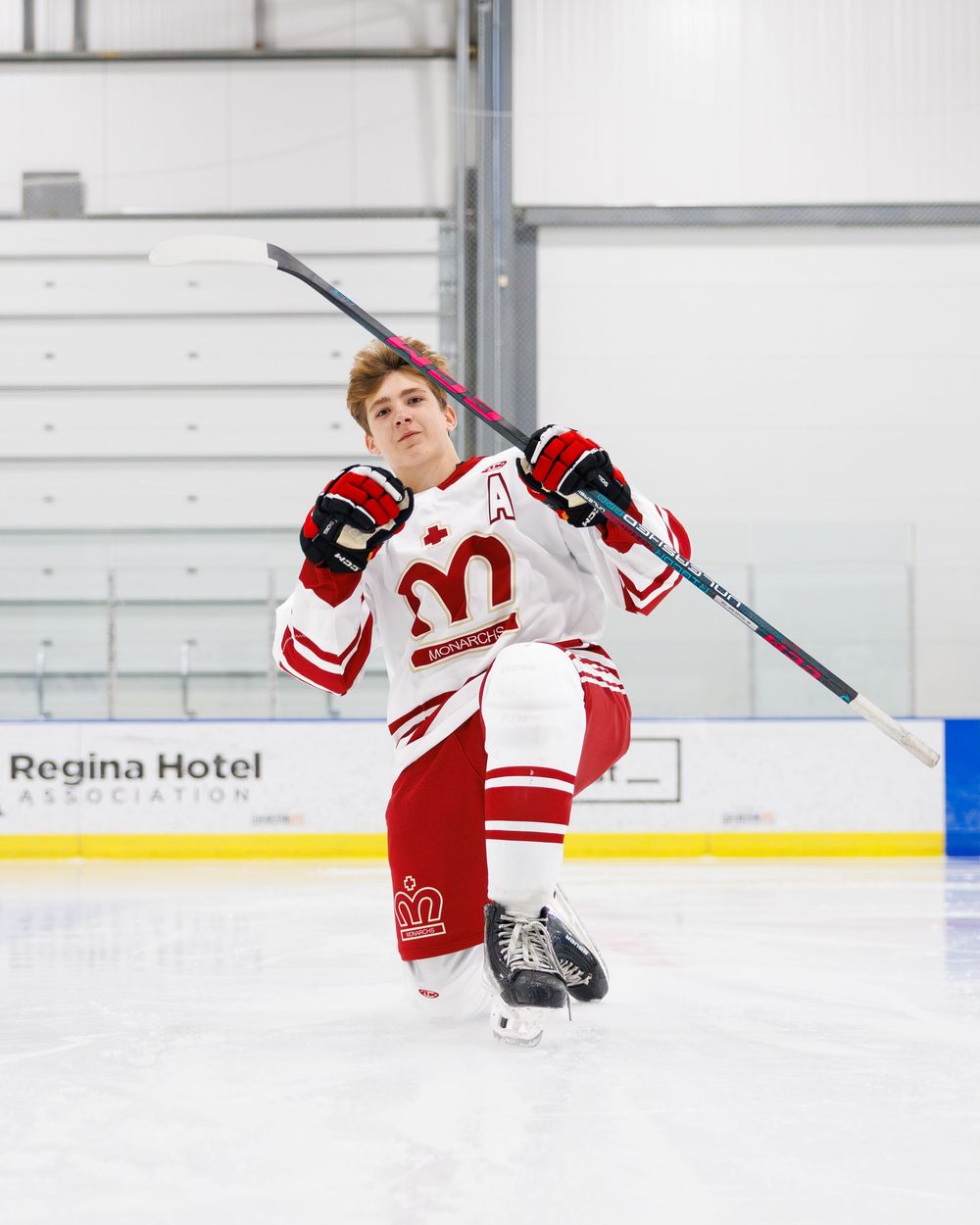 Young hockey player in red and white uniform poses on ice rink holding stick overhead at Regina Hotel Association arena.
