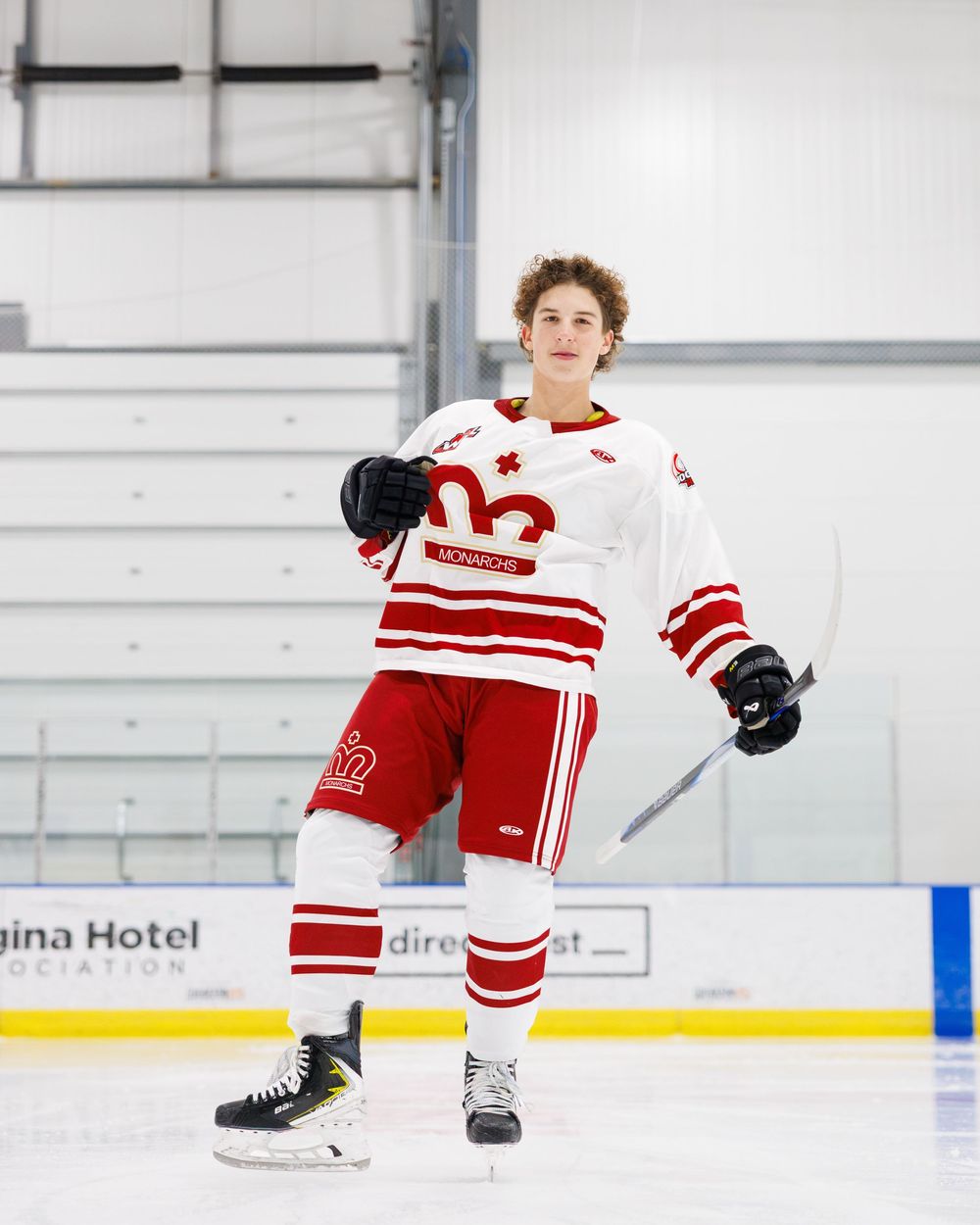 Hockey player in white and red uniform holding a stick and helmet on an indoor ice rink.