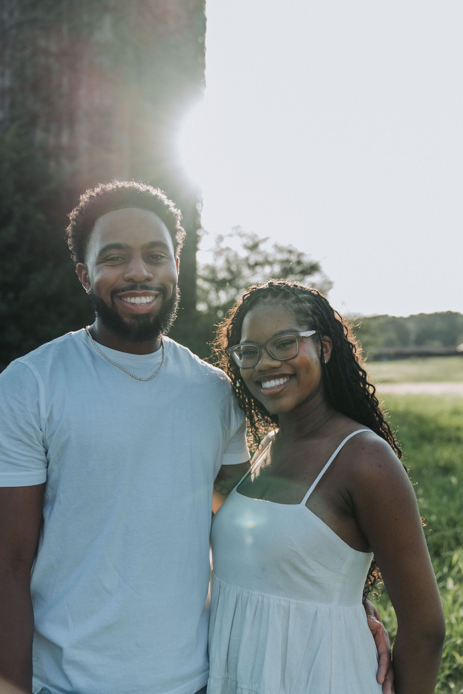 A smiling young Black couple poses outdoors in casual summer attire, bathed in warm golden sunlight near lush greenery.