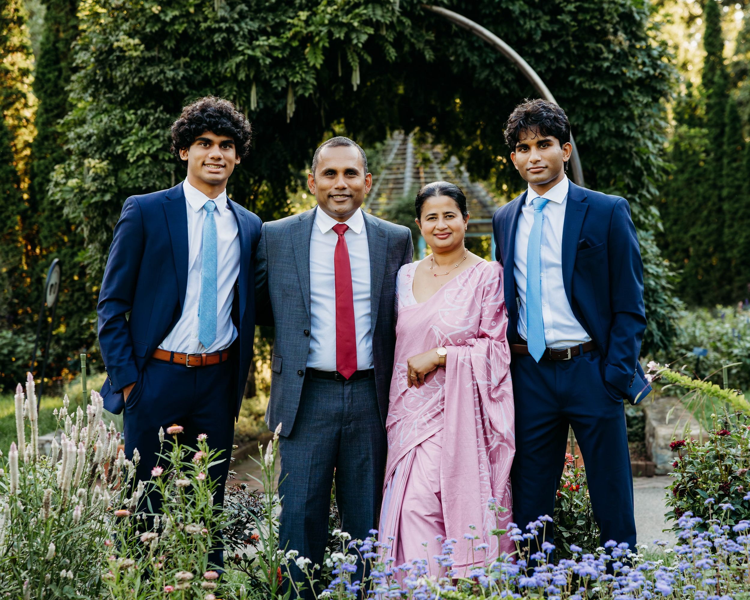 Family of four posing in a garden, two young men and a woman in pink saree flanking a man in suit with red tie.