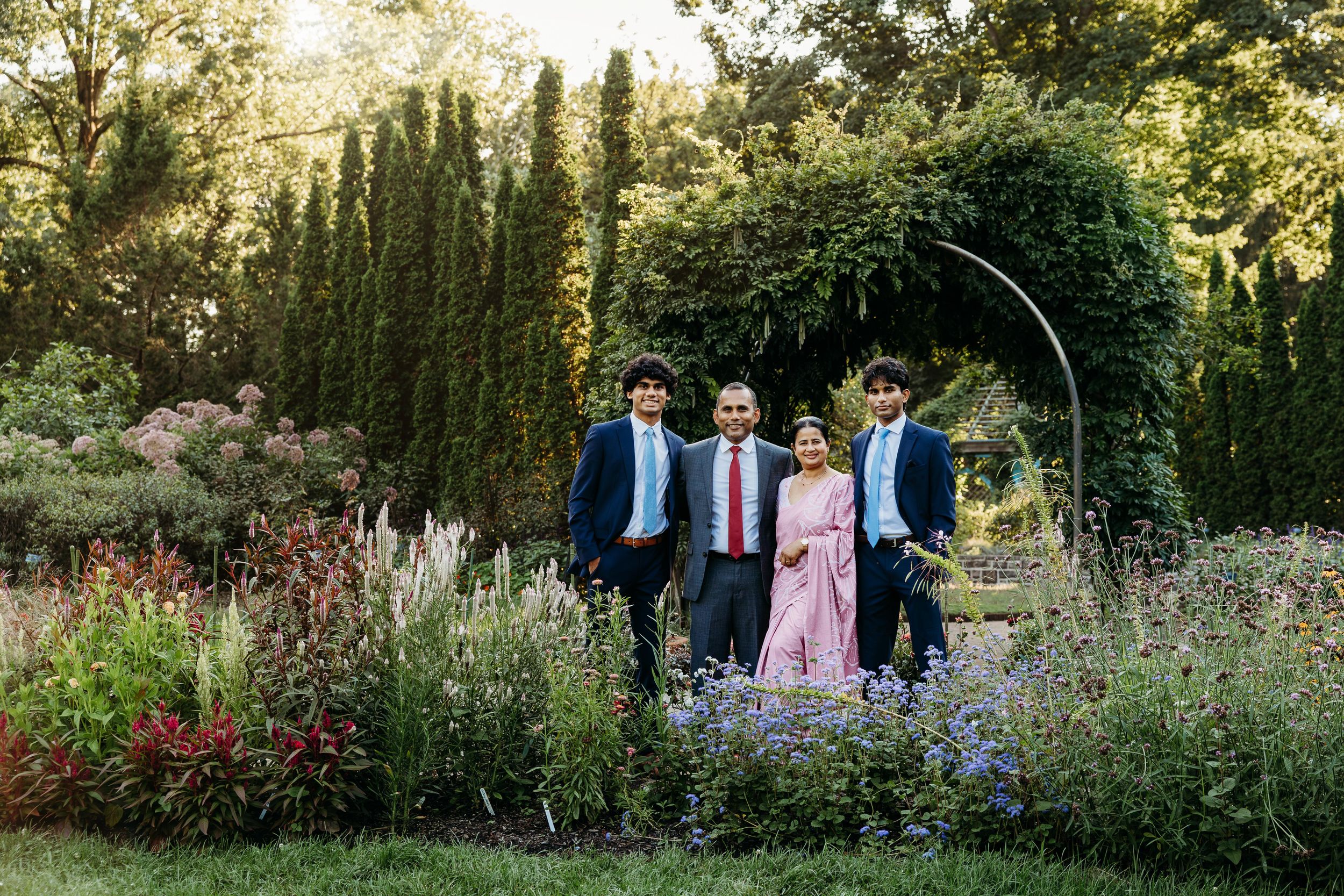 Family of four posing in a lush garden with tall cypress trees and colorful flowers under a floral arch.