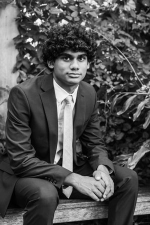 Young man in suit and tie sitting on bench with curly hair, black and white portrait photo outdoors.