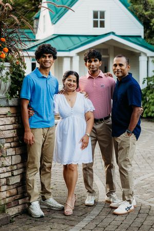A smiling family of four poses outdoors in front of a white building with a teal roof on a brick pathway.
