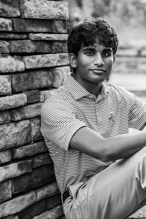Black and white portrait of a young man in striped polo shirt leaning against a stone brick wall outdoors.
