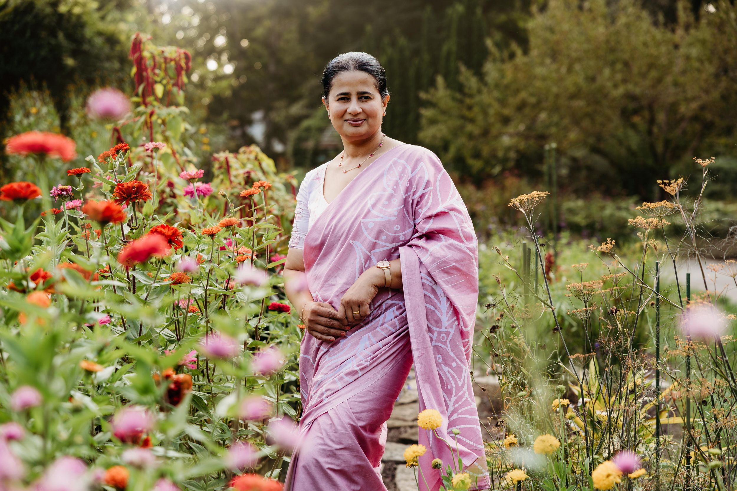 Woman in pink saree standing among colorful garden flowers, smiling outdoors in lush green surroundings.
