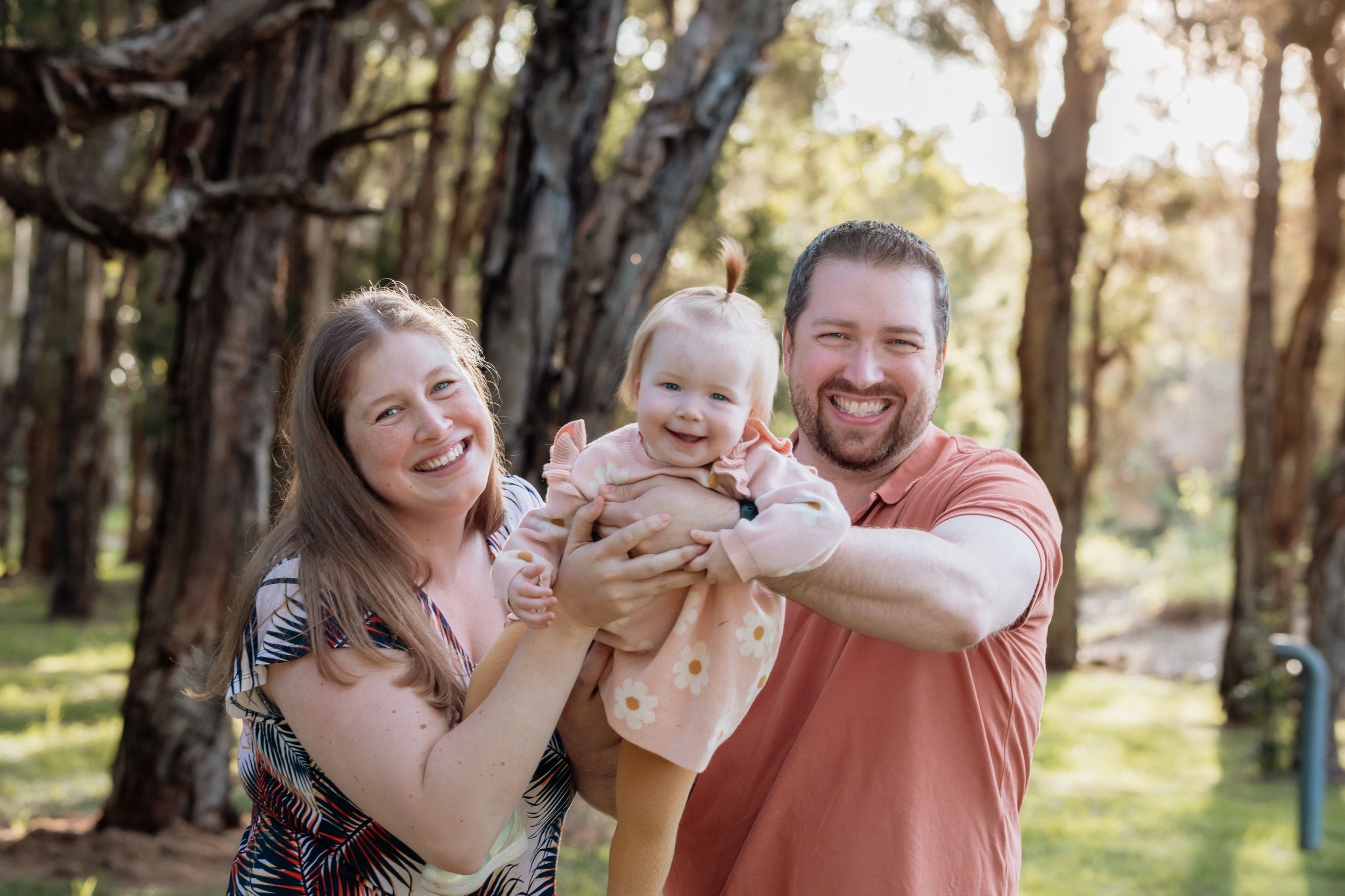 Happy family with smiling parents holding a laughing baby outdoors in a sunlit park with trees during family photoshoot in Beaumont Hills.