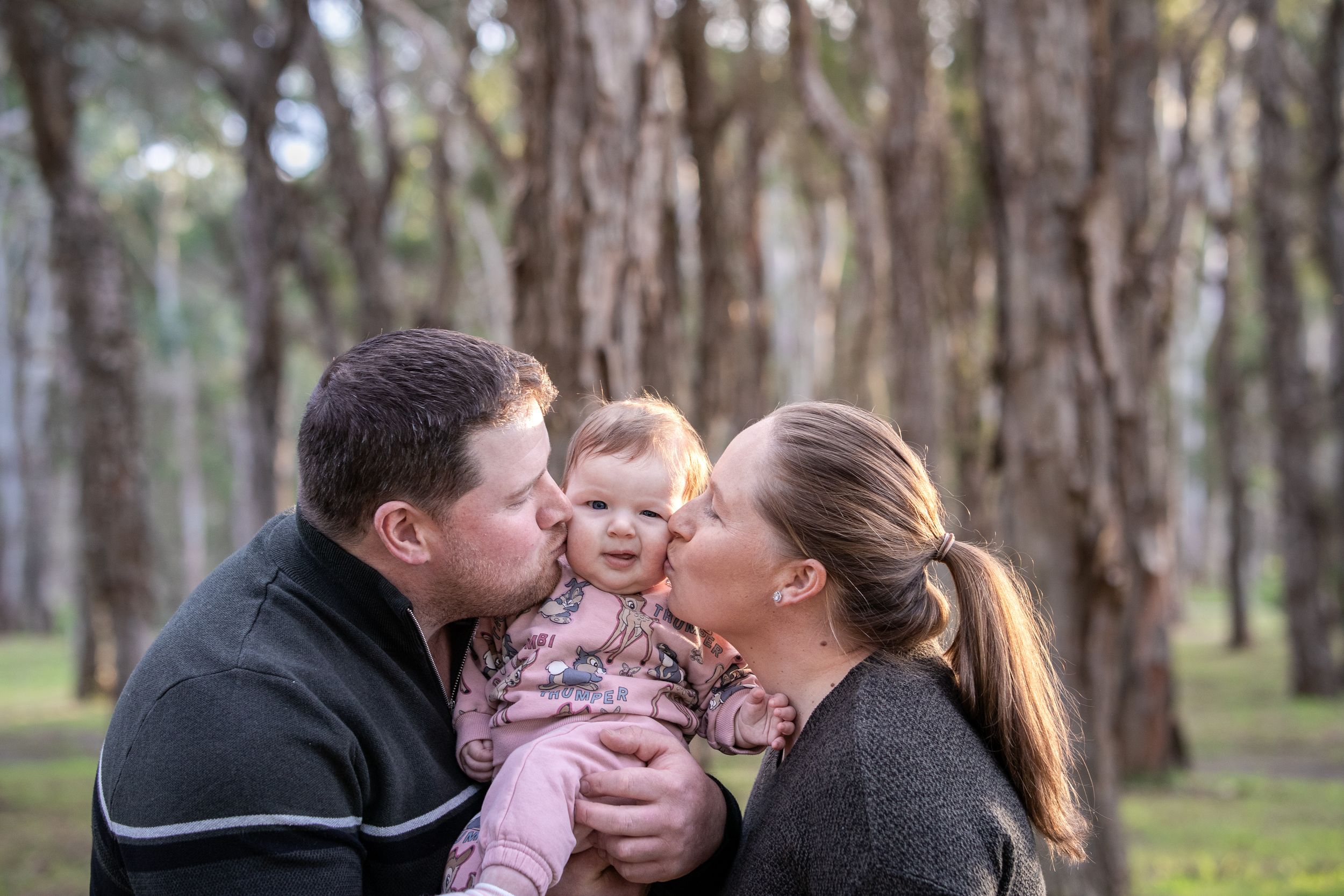 Parents kissing their baby girl on the cheeks in a lush green forest in Beaumont Hills during a family photo session.