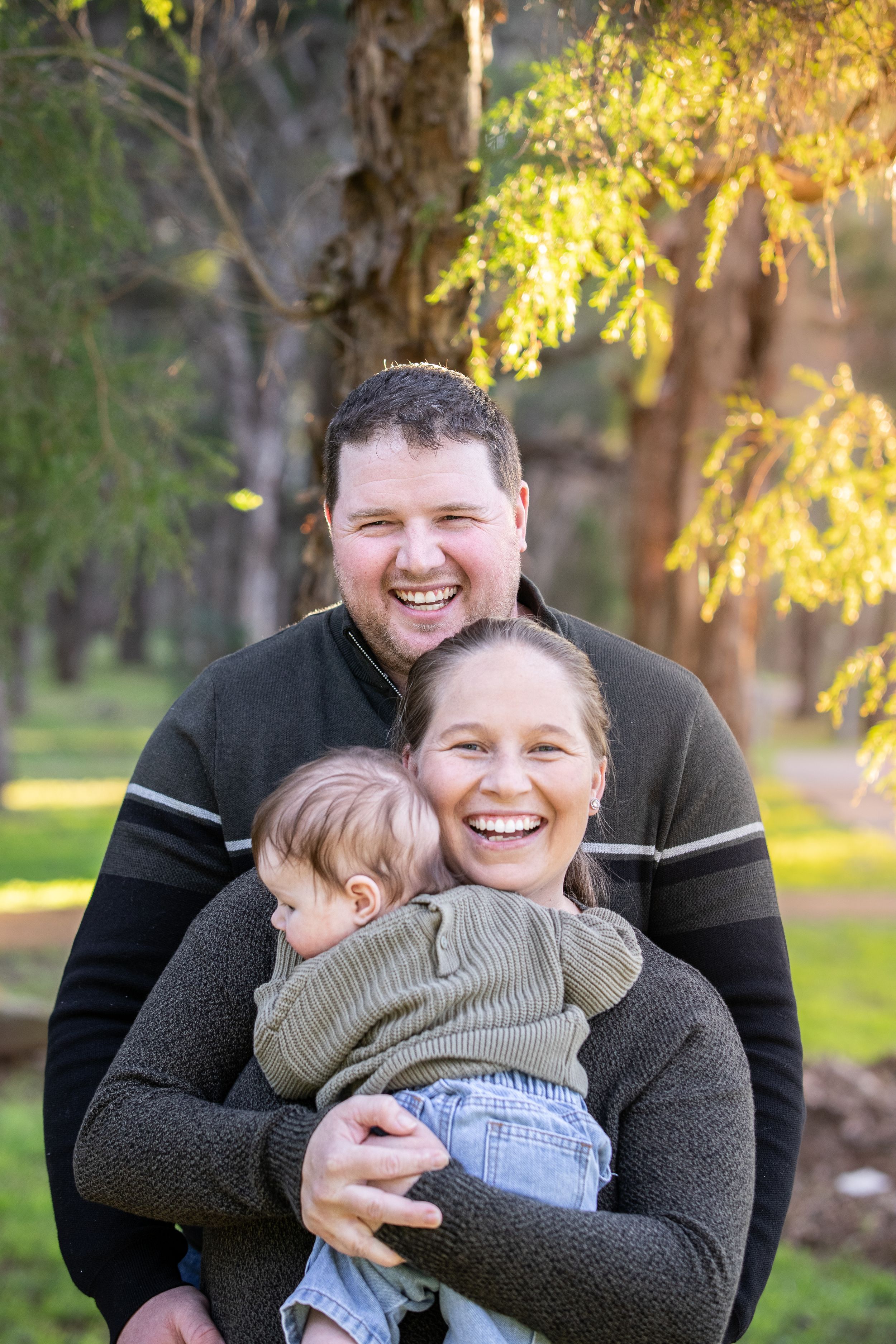 Happy family portrait outdoors, parents smiling and holding baby in green park setting with golden sunlit trees in Beaumont Hills.