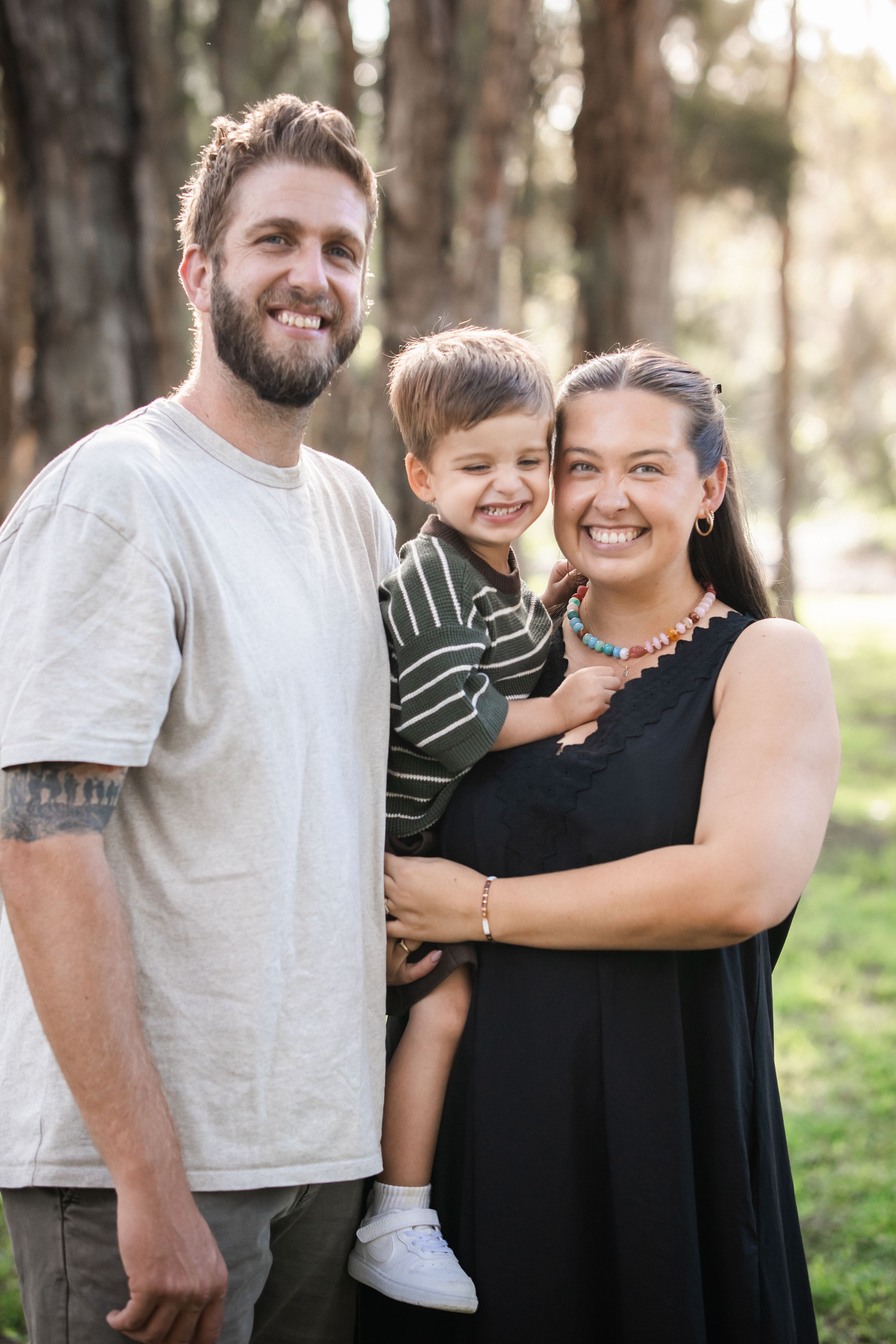 Happy family of three smiling outdoors in a park in Beaumont Hills, parents holding young toddler boy between them during family photoshoot.