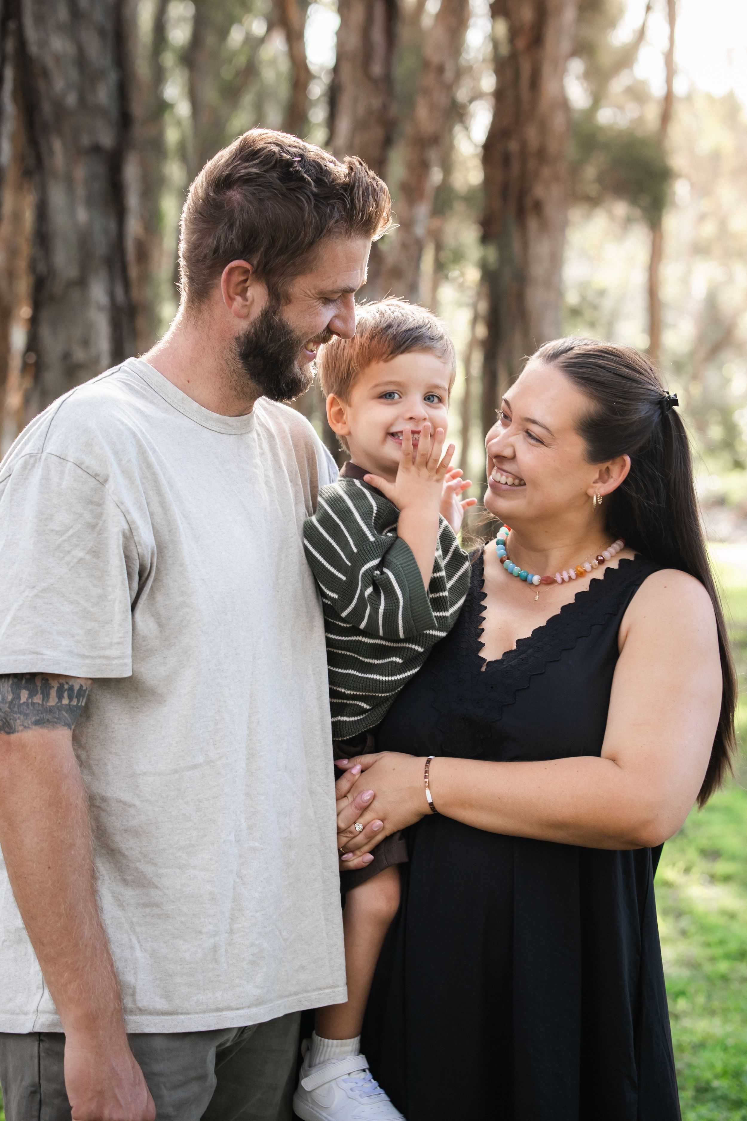 Happy family of three outdoors, mother in black dress, father holding toddler son, smiling together in park in Beaumont Hills.