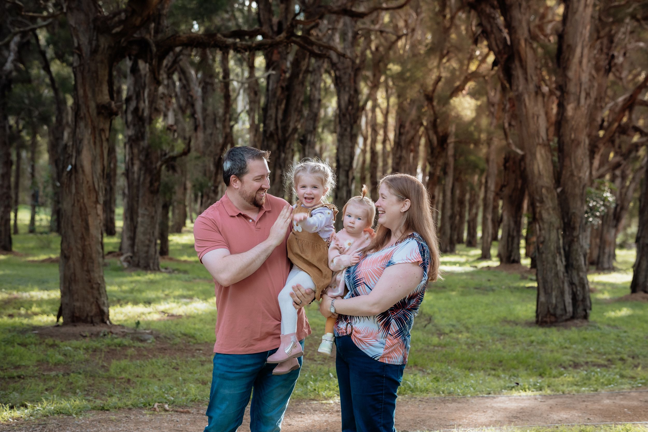 Family of four smiling outdoors in a paperbark tree-lined park in Beaumont hills, parents holding two young toddlers on a sunny day.
