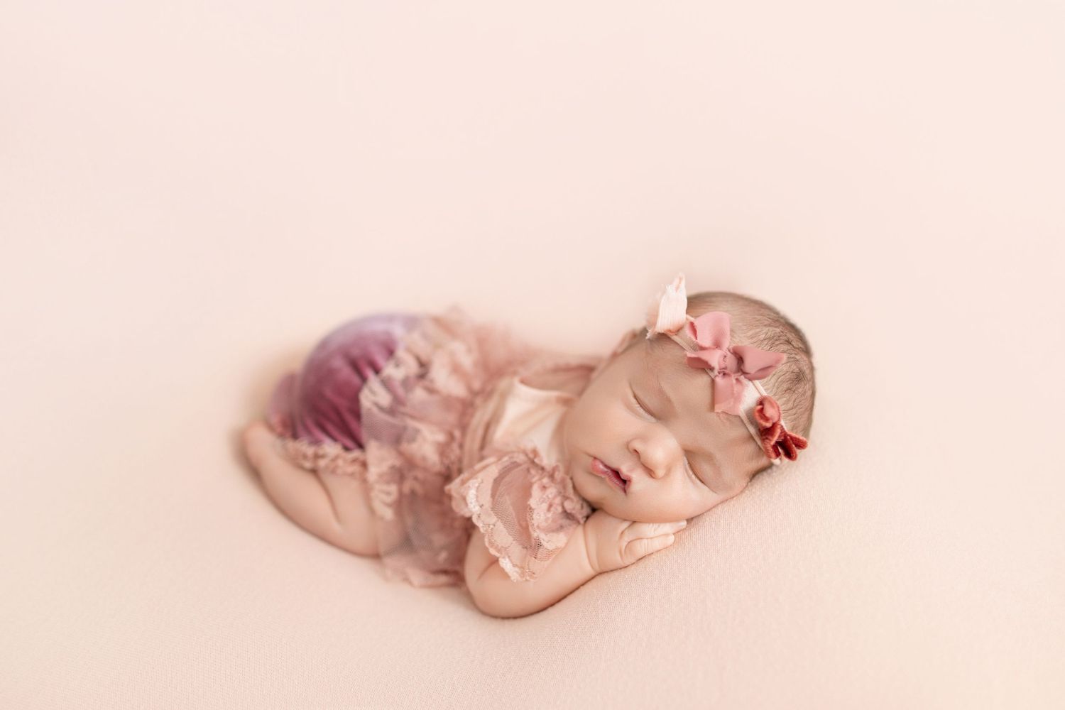 Sleeping newborn baby girl in pink lace outfit and bow headband on soft peach background.