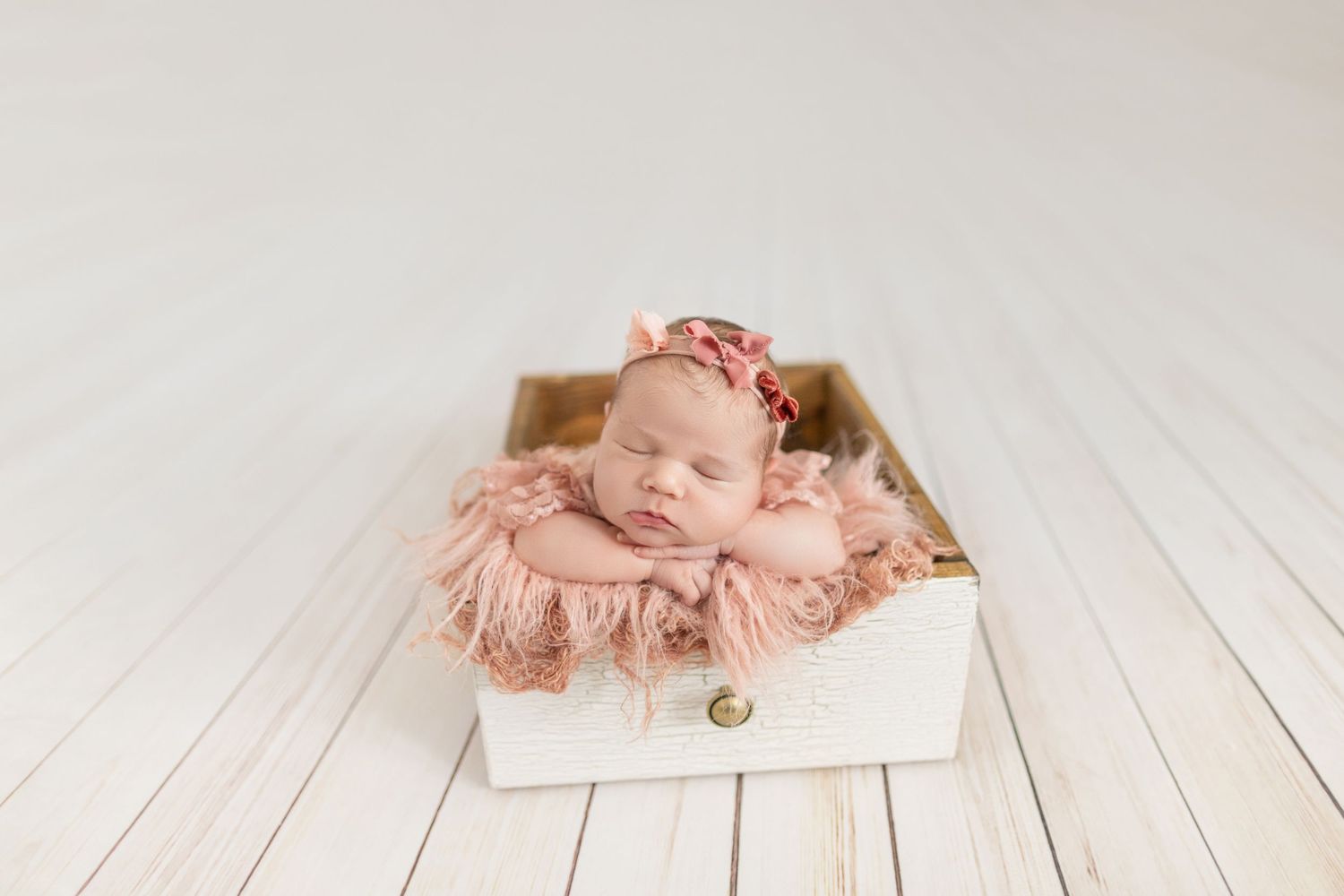 Newborn baby girl sleeping peacefully in a white wooden box with pink lace blanket and floral headband.