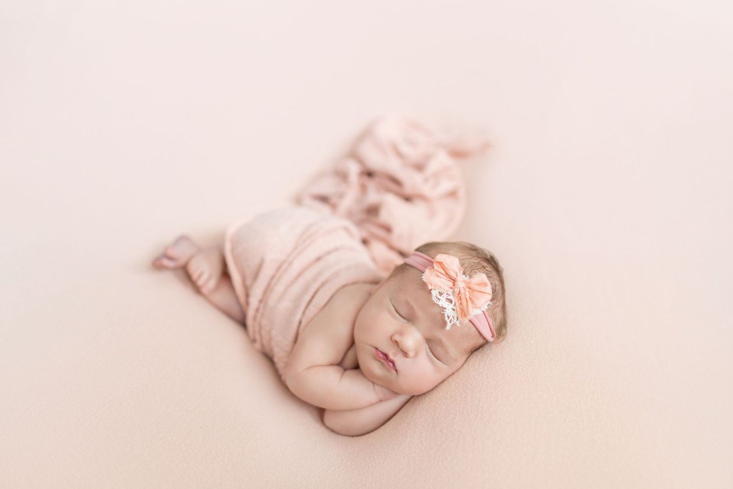 Sleeping newborn baby girl wrapped in soft pink fabric with floral headband on cream background.