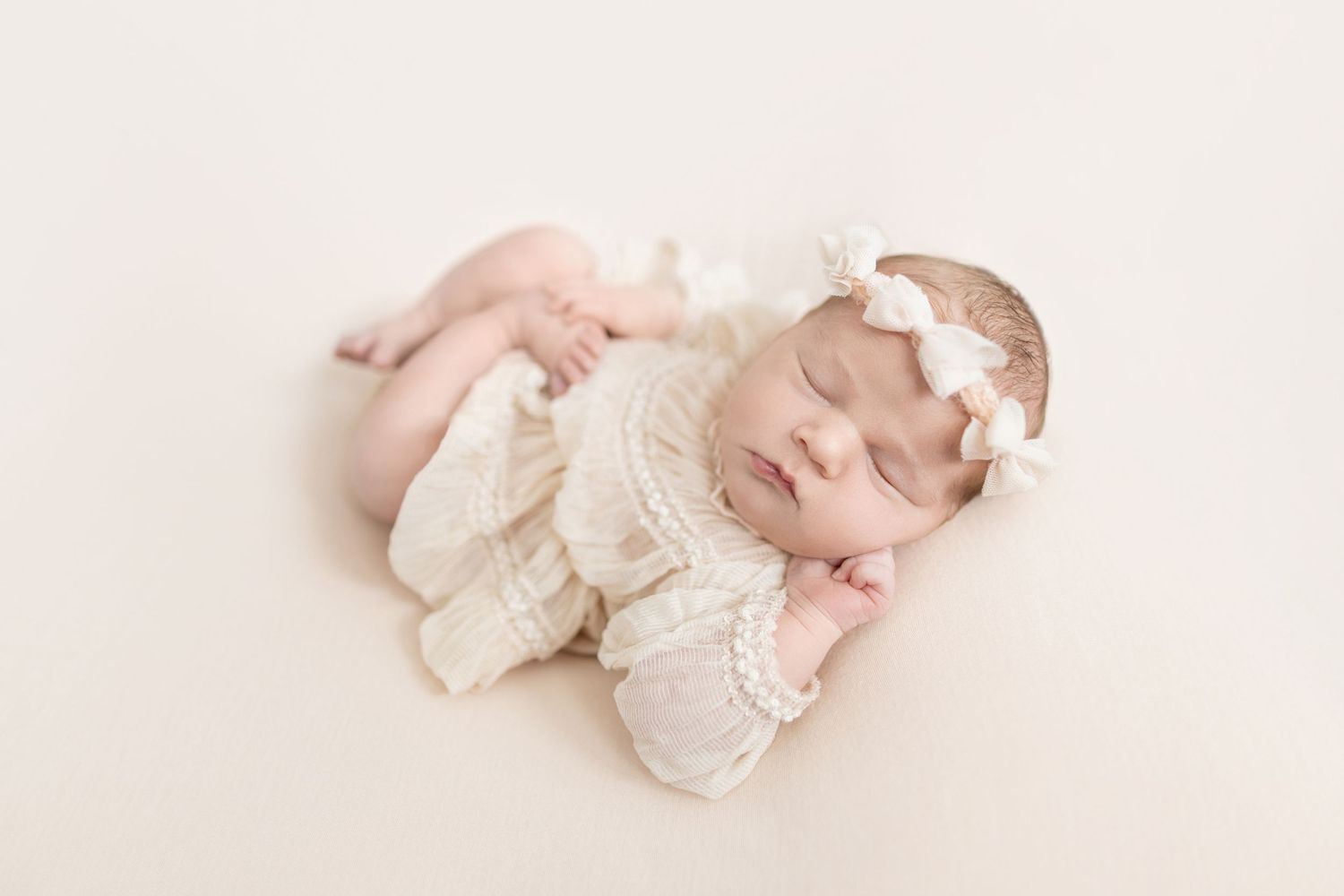 Sleeping newborn baby girl in cream knit romper and bow headband posed on white backdrop.